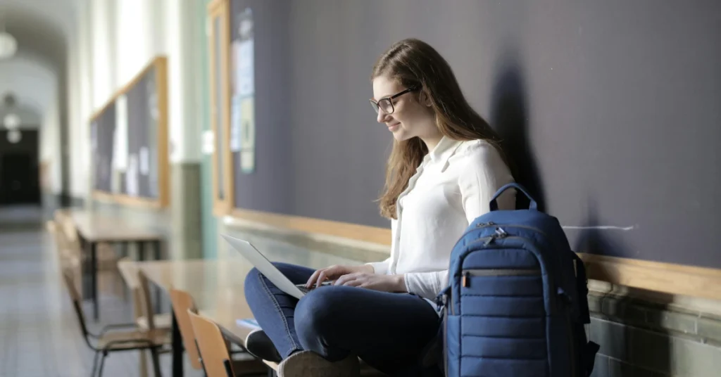 Student Using Laptop While Sitting on Table Near Blue Backpack